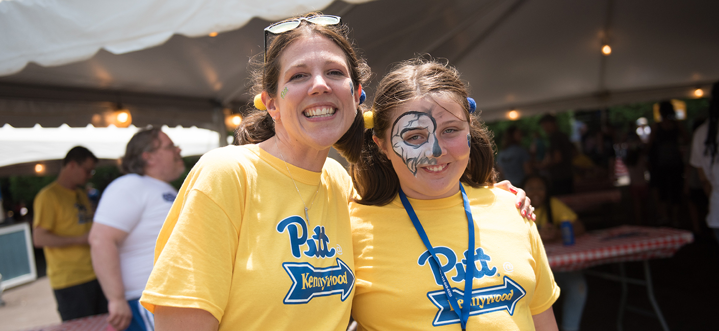 Amy Kleebank with her daughter, Amelia, at Staff Council's Pitt Day at Kennywood, wearing bright yellow Pitt shirts. Amelia has face painting on her right eye.