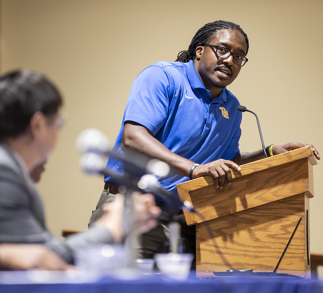 Ron Idoko in a blue collared shirt at a lectern, speaking into a microphone.