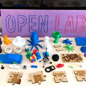 A purple sign that reads, "Open Lab," on a table filled with 3D-printed objects, in different colors.