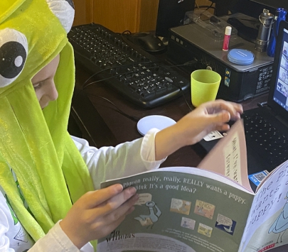 Child wearing a green alien hat reads a book to people on a Zoom virtual call
